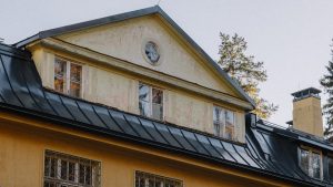 Close-up of a vintage house with a rustic yellow facade and black metal roof in an autumn setting.